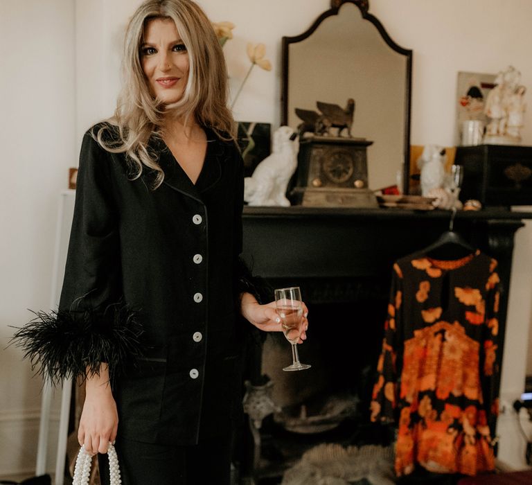 Bride in black fur trimmed pyjamas holding speaking wine and small beaded tote bag smiles as she gets ready for her wedding