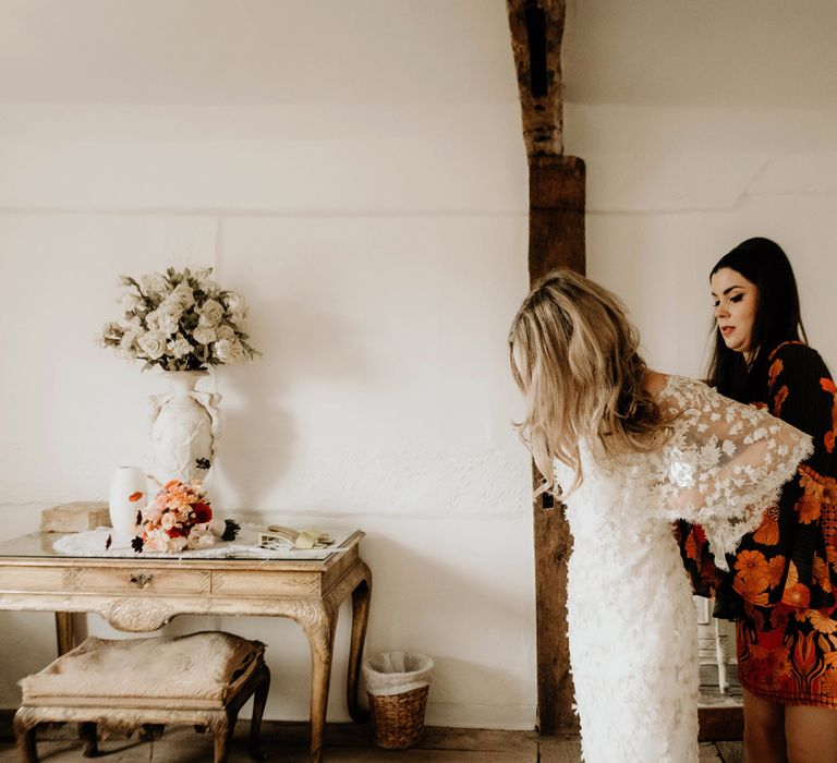 Bridesmaid in black and orange print floral dress helps bride in long sleeve appliqué wedding dress to do up her dress