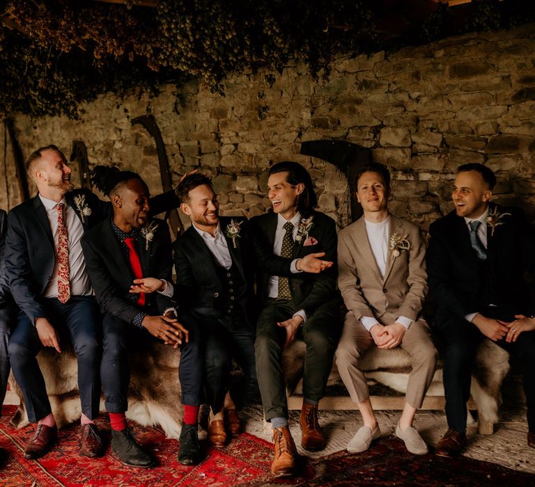 Groom in dark three piece suit sits with groomsmen in mixed groomsmen suits on wooden bench in stone barn for rustic wedding