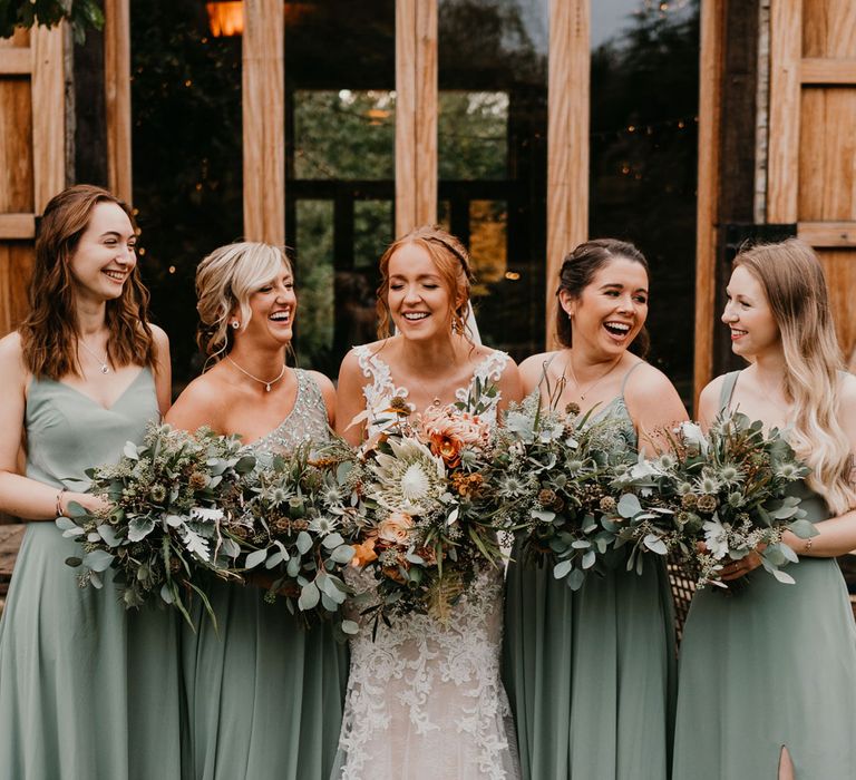 Bride with braided updo wearing lace wedding dress holding mixed bridal bouquet stands with bridesmaids in mismatched sage bridesmaid dresses holding green bouquets 