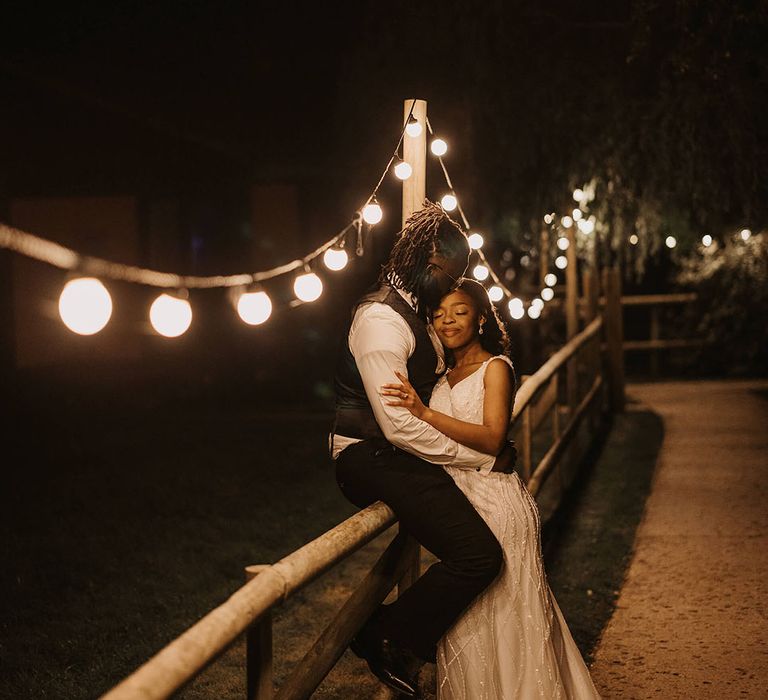 Bride & groom stand on bridge in front of festoon lighting