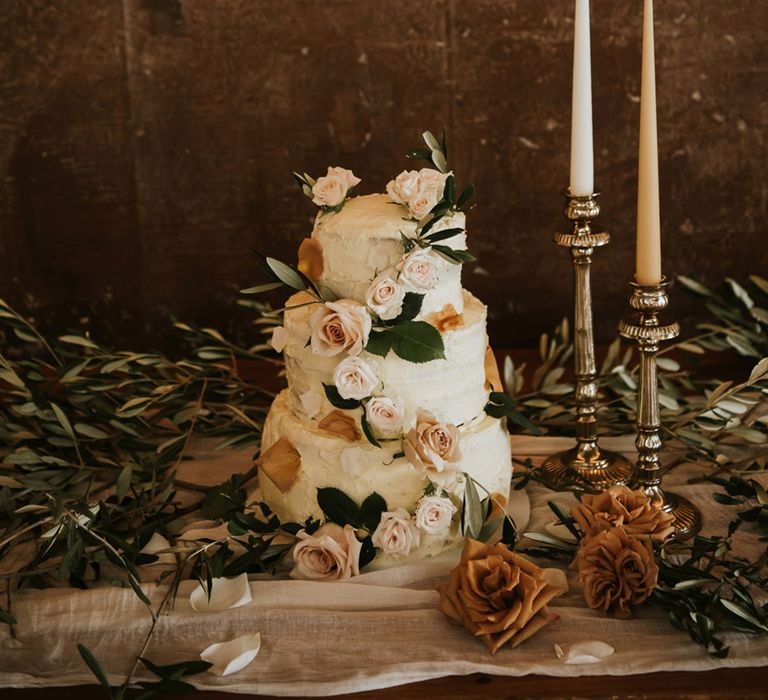 Three tier buttercream wedding cake decorated with blush roses on a cake table with candlesticks and taper candles 