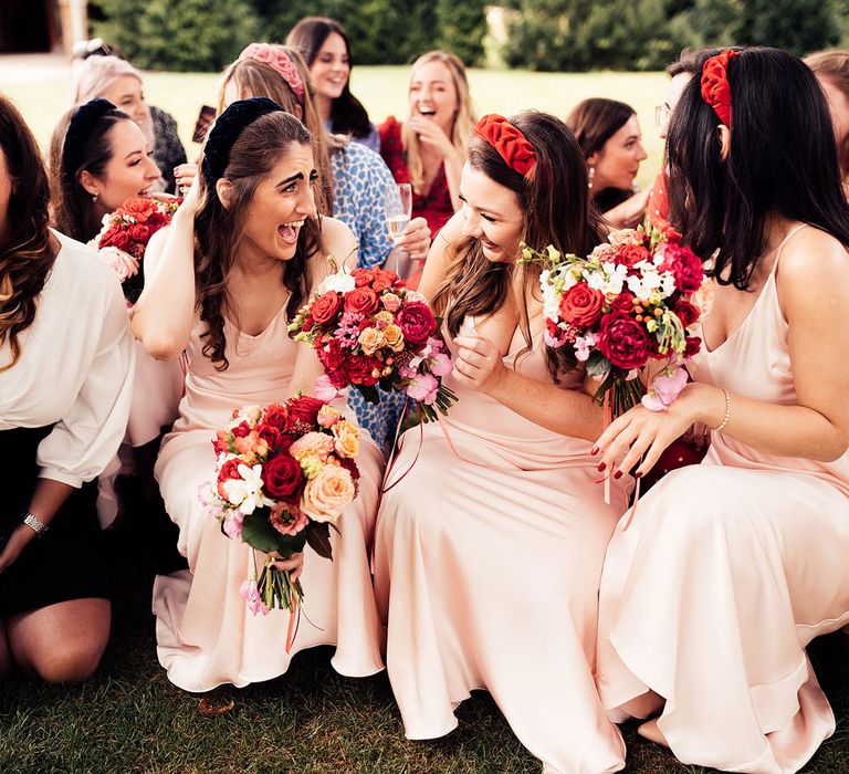 Bridesmaids in baby pink satin bridesmaid dresses holding mixed bouquets and wearing bespoke bridesmaid headbands laugh during sugar ceremony