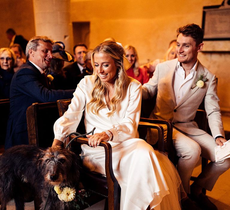 Bride in satin long sleeve wrap top and veil wearing silver heeled sandals sits next to groom in light grey suit during church wedding ceremony