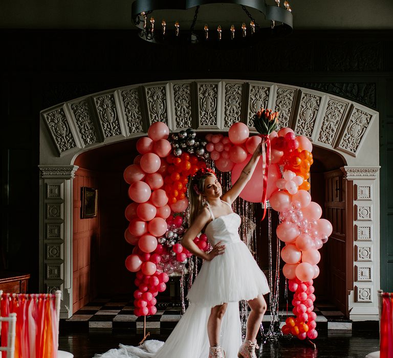 Bride in a short wedding dress wearing roller skates holding a tulip bouquet in the air at her fun, retro wedding with balloon installation