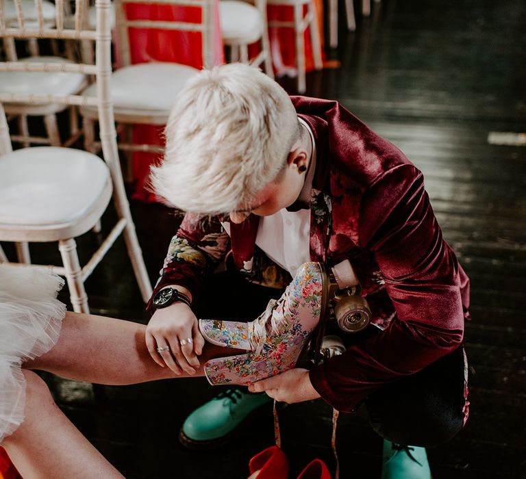 Bride with short platinum blonde hair and a burgundy velvet jacket putting on her brides floral retro roller skates 