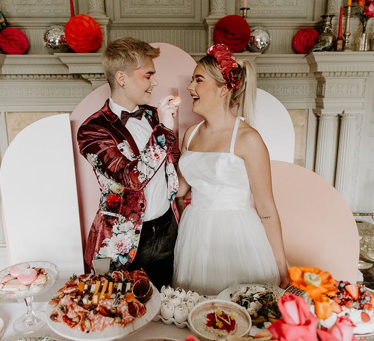 Two brides at the food table with pink geometric backdrop, velvet jacket and tulle skirt wedding dress 