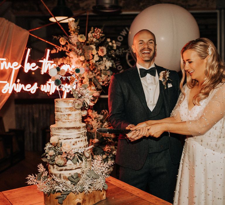 Groom in navy tweed suit with bow tie and buttonhole stands with bride in white wedding dress with pearl mesh overlay as she cuts three tier semi-naked wedding cake