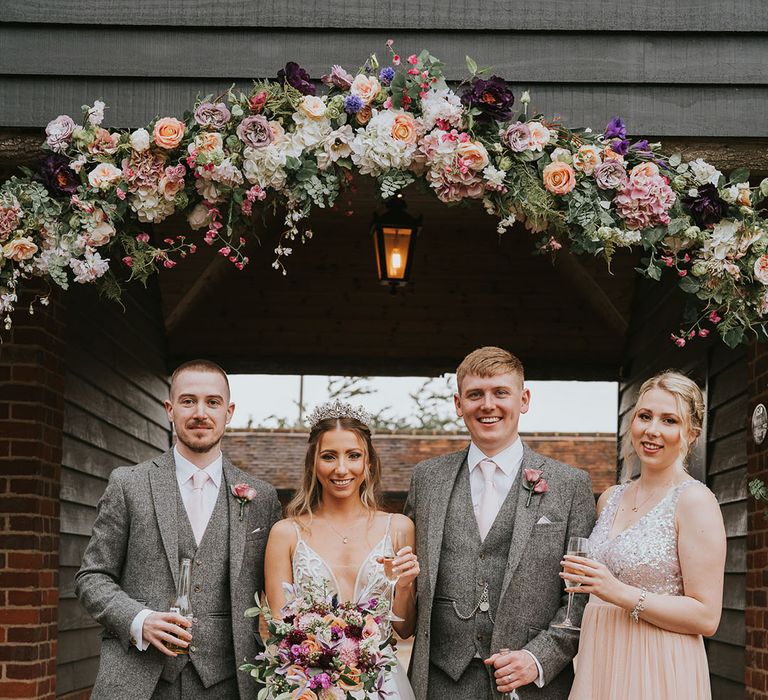 Bride and groom smile with bridesmaid and groomsman under arch of flowers of bright colours