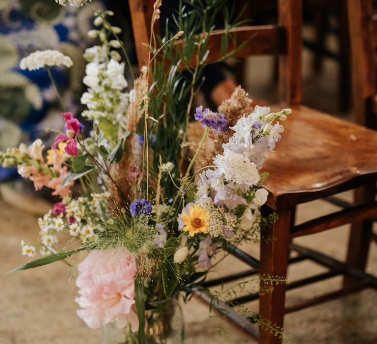 Colourful wildflower wedding flower displays in glass vases for aisle decoration next to dark wooden chairs