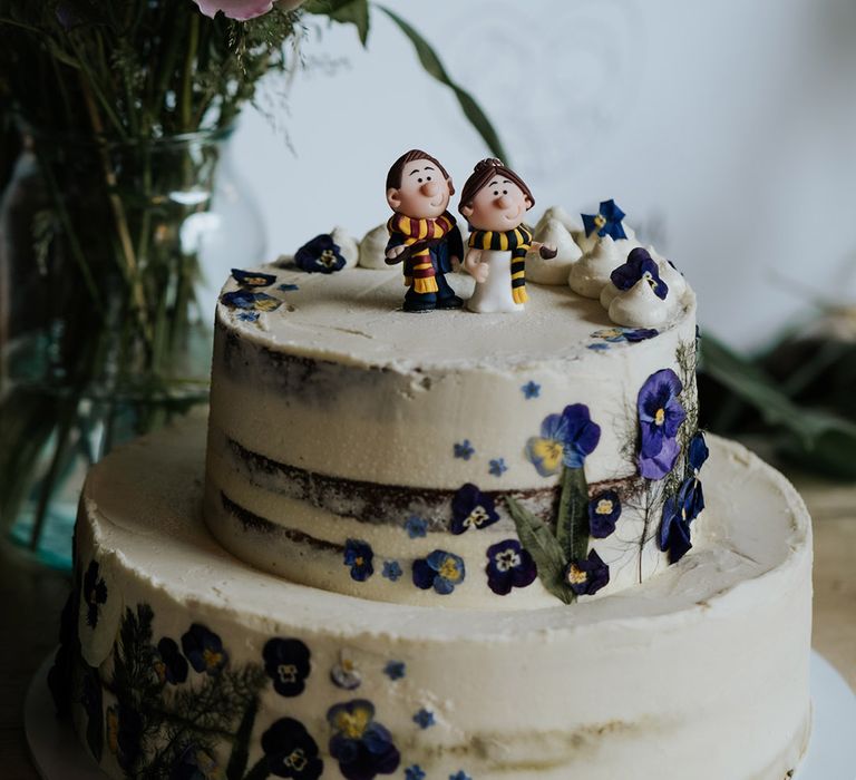 Semi-naked two-tier wedding cake with white frosting and edible flowers topped with custom bride and groom cake topper in Harry Potter style scarves 