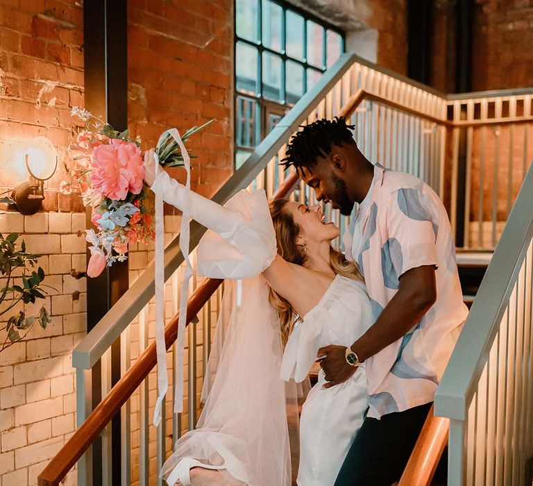 Groom in a pastel patterned shirt and Converse trainers embracing his bride in a short wedding dress with long veil holding her pink and blue bouquet in the air 