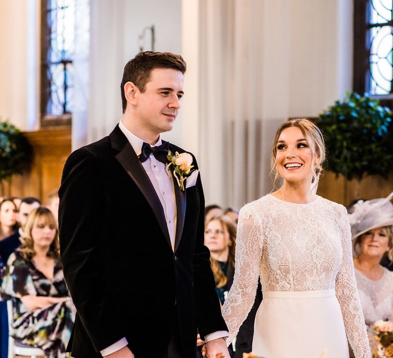 Bride in lace top long sleeve fitted wedding dress looks adoringly at groom in black tie with candles and gold decor