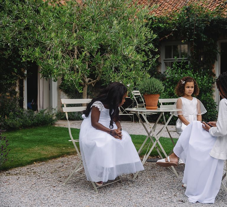 Flower girls sit together in white dresses and white shoes seated on garden furniture