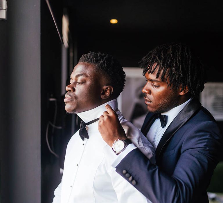 Groomsmen in blue tuxedo helps groom put on his bow tie or wedding day 