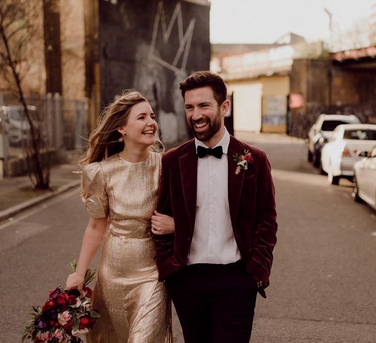 Bride in gold gown and star earrings links arms with groom in red velvet suit jacket at 100 Barrington city wedding venue