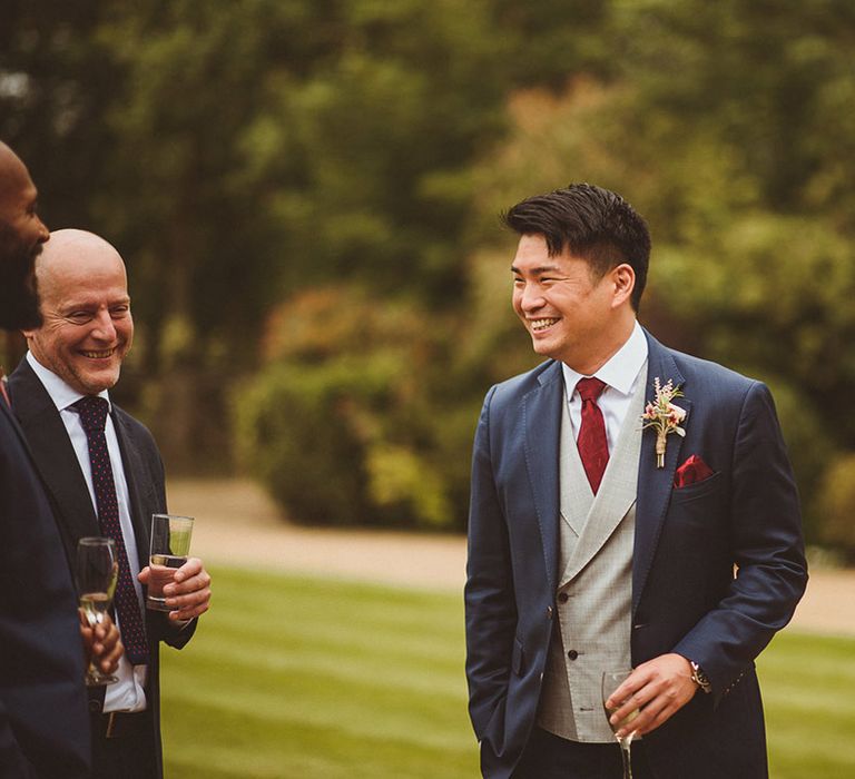 Groom smiles holding a champagne glass wearing a blue suit, red tie and grey waistcoat as he talks to guests