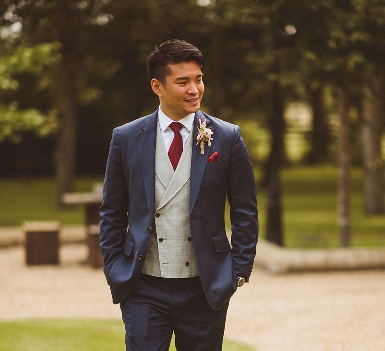 Groom in blue suit with grey waistcoat and red tie and handkerchief as he walks with his hands in his pockets