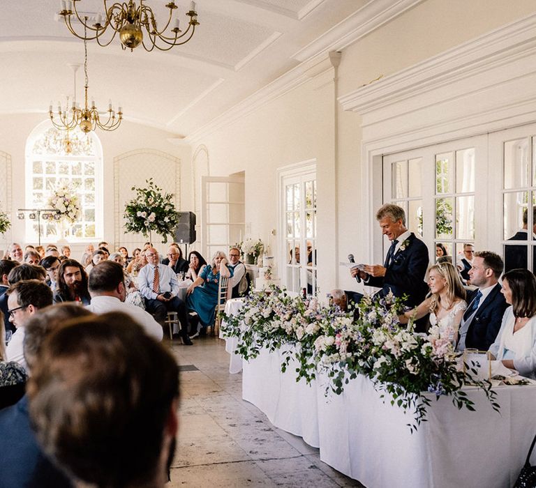 Father of the bride stands as he reads out his speech for traditional wedding with top table decorated with colourful spring flowers 