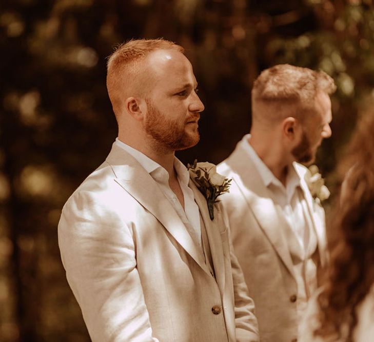 Groom wears cream three piece suit with relaxed open shirt and floral buttonhole 