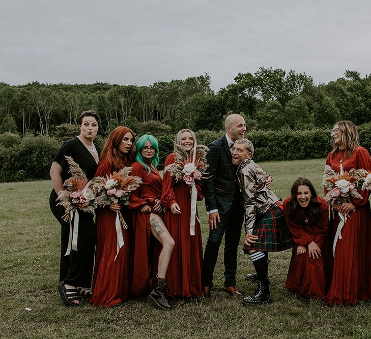 Grooms stand beside their bridesmaids who wear long sleeve red dresses complete with homemade floral bouquets 