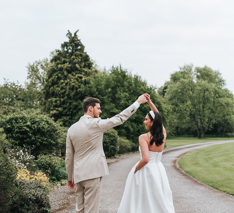 Groom in a neutral wedding suit spins the bride around in her strapless pleated wedding dress 