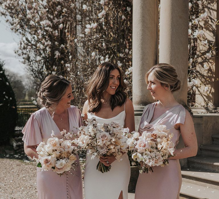 Bride in white satin wedding dress with bridesmaids in pink satin dresses all holding matching neutral and blush pink bouquets 