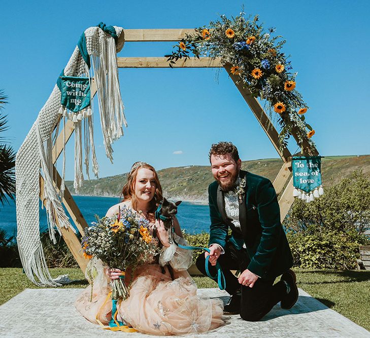 Bride & groom kneel with their dog in front of DIY wedding arch adorned with sunflowers and green foliage alongside blue fabric wedding banners