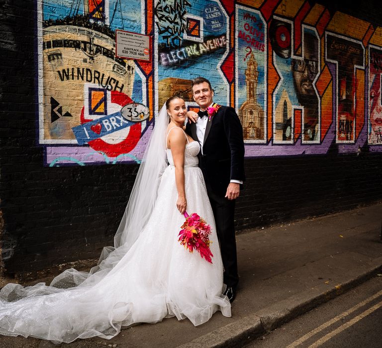 Bride holding bright floral bouquet with pink pampas grass and her groom in black tie stand in front of colourful graffiti wall in Brixton