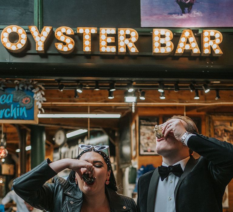 Bride wears leather jacket and holds a glass of champagne as her and her groom eat oysters in Borough Market