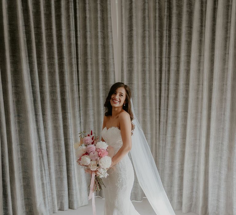 Bride wears strapless lace fitted Pronovias wedding dress with floor-length veil and holds white and pink floral bridal bouquet