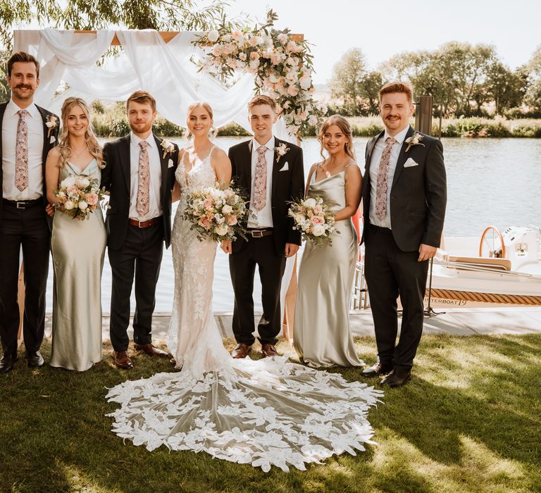 Bridesmaids wearing pastel green satin bridesmaid dresses stand beside groomsmen wearing black suits with white shirts and floral ties in front of the Thames