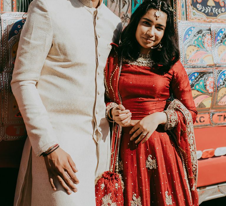 Bride wears gold accessories and beautifully embellished red bridal separate as she stands beside her groom outdoors  