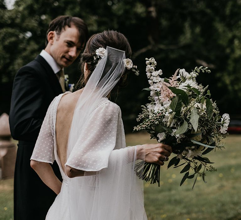 Bride in batwing sleeve lace detail wedding dress with open back and neutral toned wedding bouquet