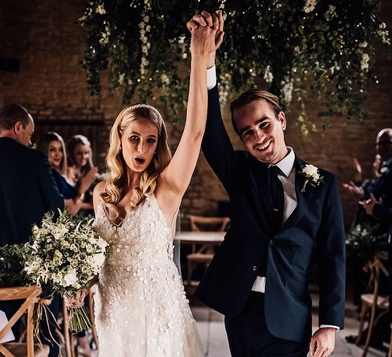 Groom in a navy blue suit with a white rose buttonhole walks back down the aisle with the bride 