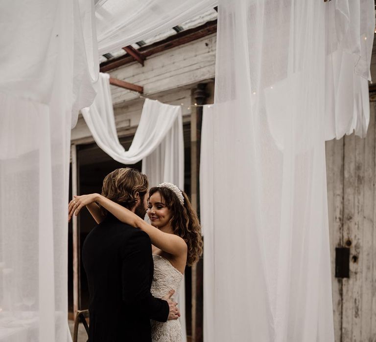 Bride in sleeveless lace wedding dress with silver belt and pearl headband and groom in black tux dancing 