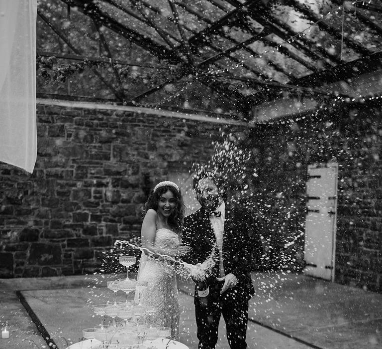 Bride in sleeveless lace and pearl wedding dress and pearl headband with groom in black tux doing Champagne pop 