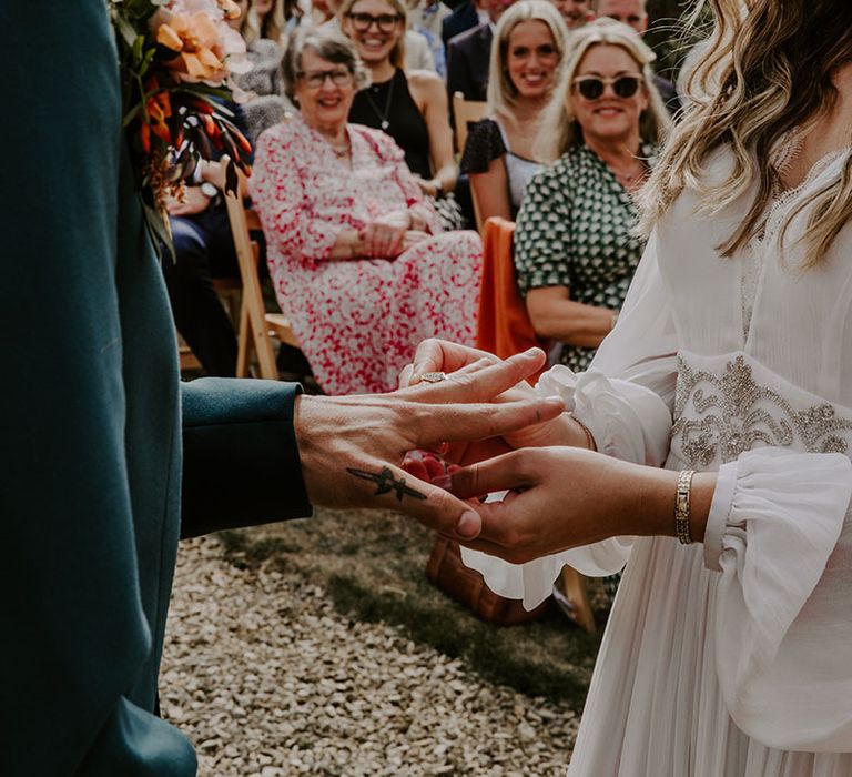 Bride places ring on her grooms finger as they stand outdoors during wedding ceremony 