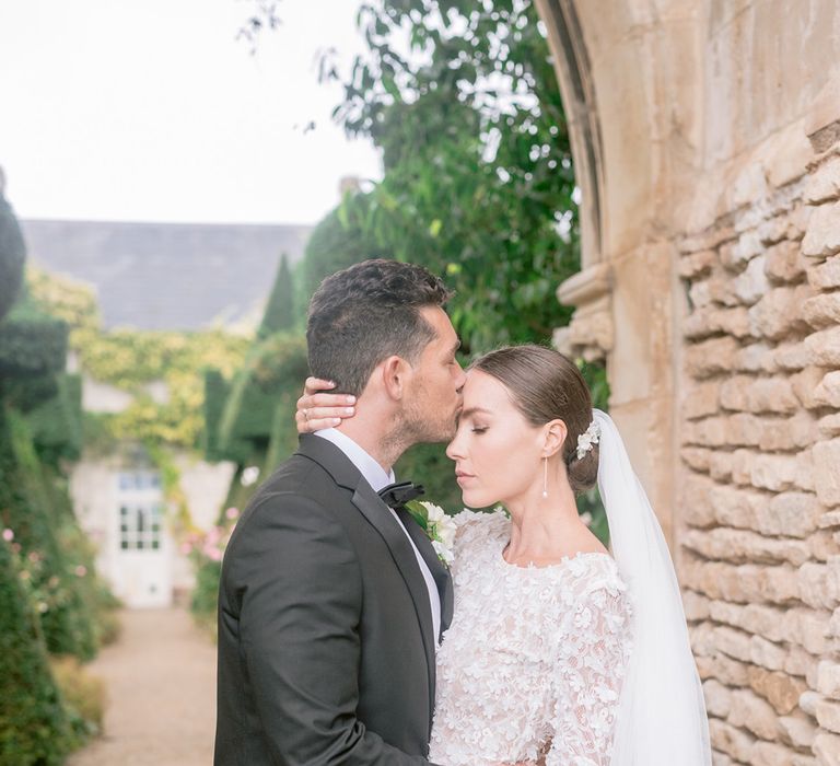 The groom in black tie kisses the bride in a flower applique lace wedding dress on the forehead 