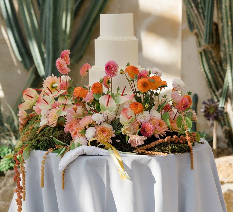 Brightly coloured floral arrangement surrounds four tier white frosted wedding cake 