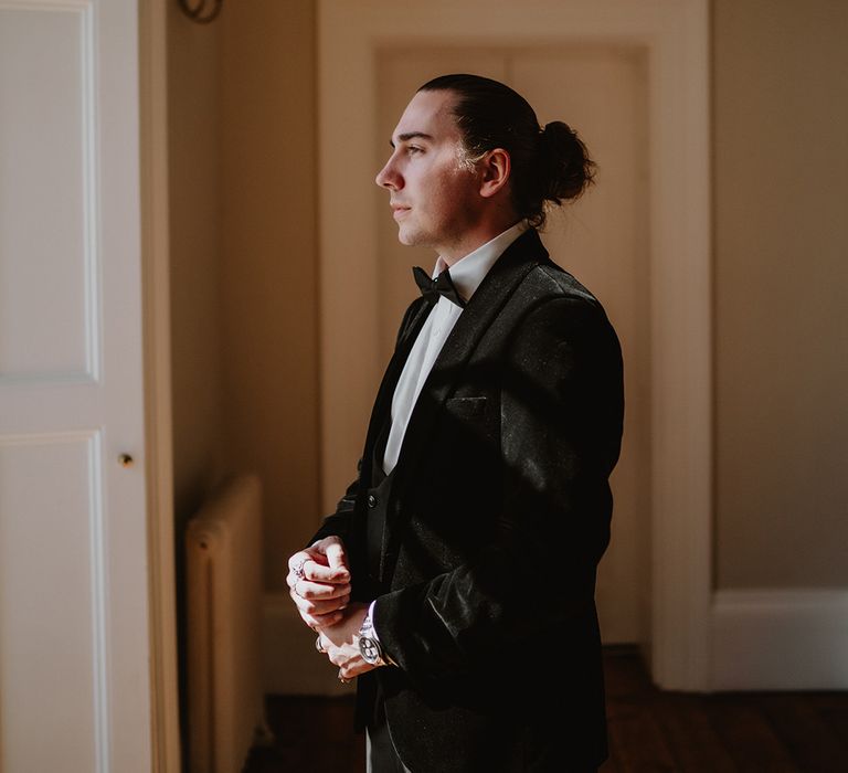 Groom in velvet-look black tux with black bowtie wearing lots of rings adjusting cufflinks at the window of St Tewdrics House