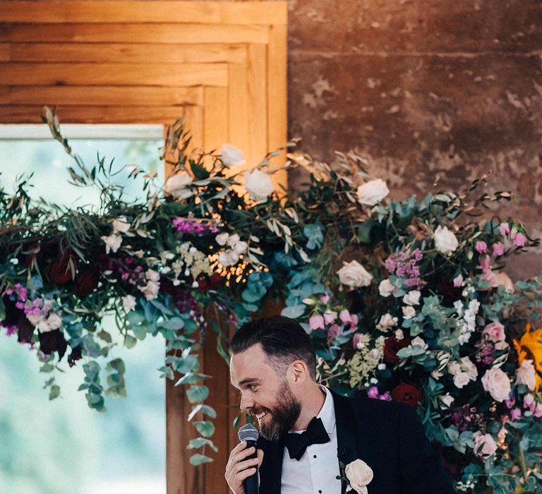 The best man gives a speech as he stands in black tie with the rose flower arch in the background 