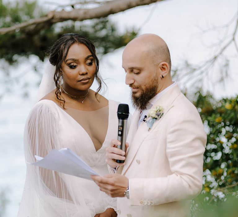 Groom in cream suit reads his vows whilst Bride in sheer-sleeved wedding dress looks at him lovingly