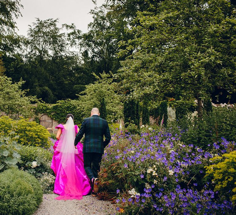 Bride in off the shoulder Vivienne Westwood front ruching pink wedding dress with two layer white and pink ombre veil and groom in dark forest green tartan suit with patterned tie, white pocket square and pink and green boutonniere walking through Middleton Lodge gardens