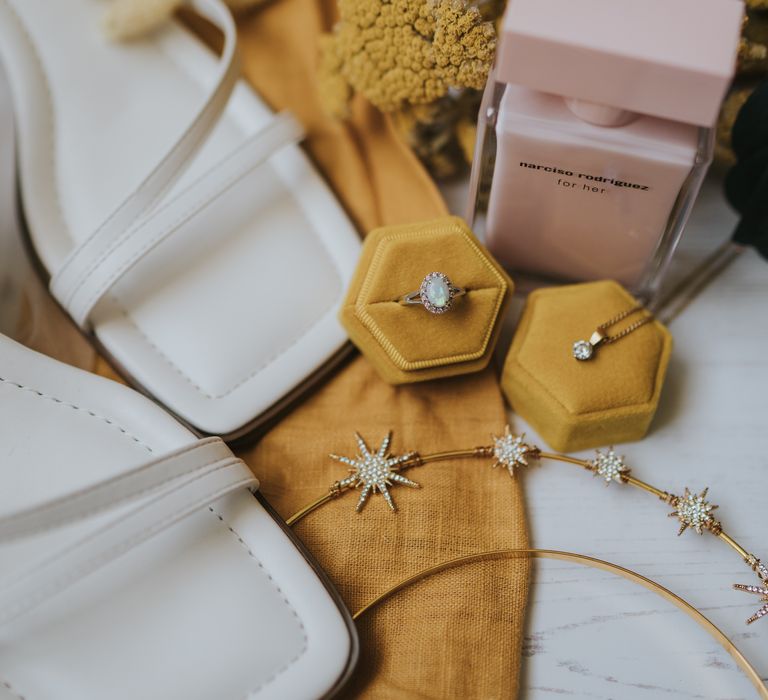 White strappy open-toe wedding heels on mustard fabric with gold headband with sparkly stars, mustard coloured ring holders, wedding rings and a pink perfume bottle 