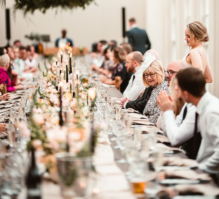 Wedding guests sitting at wedding reception room tablescape with neutral off-white table runners, rose and foliage floral arrangements, pink and black tapered candles, black candlestick holders and suspended foliage decorations