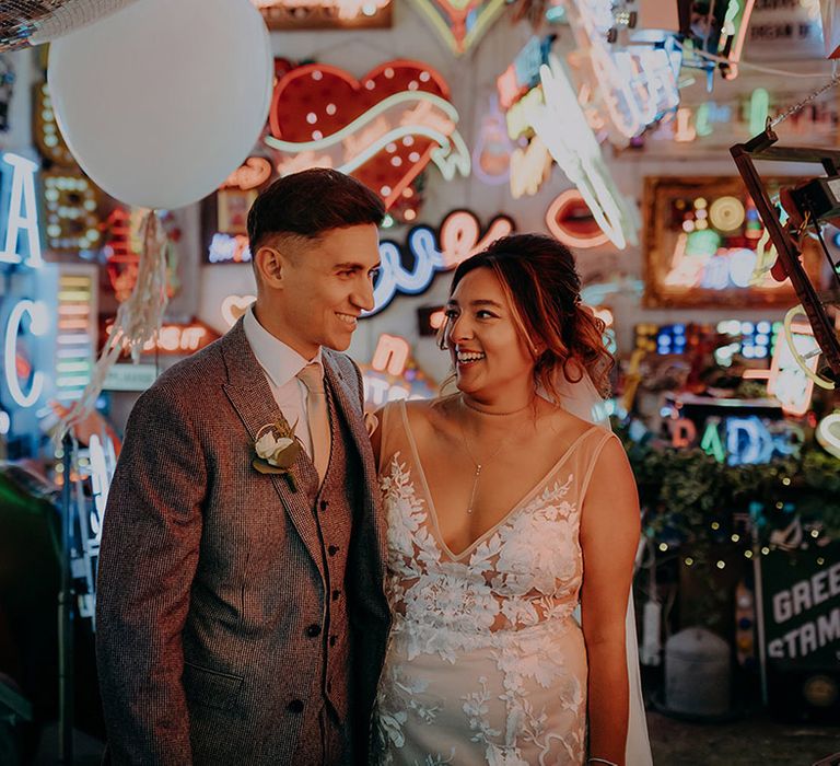 Bride in sleeveless plunge lace wedding dress and cathedral length embroidered veil and groom in grey three piece wedding suit with yellow suit and yellow pocket square standing in Gods Own Junkyard with wedding disco ball decor, neon signs and LED signs 