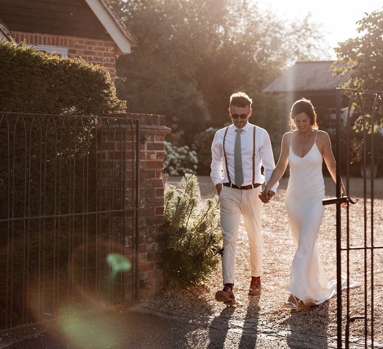 Groom in white shirt and sage green tie holds his brides hand as they walk outdoors during golden hour