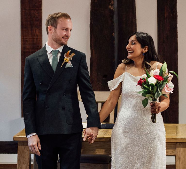 Indian bride in sequin embellished wedding dress looks lovingly toward her groom in suit and floral buttonhole 