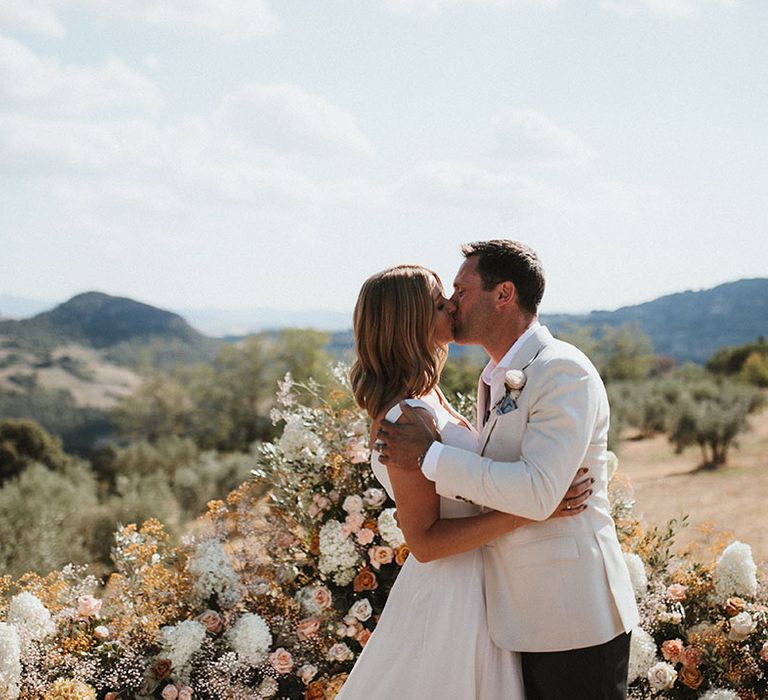 Groom kisses his bride in front of wedding altar flowers in crown shape 